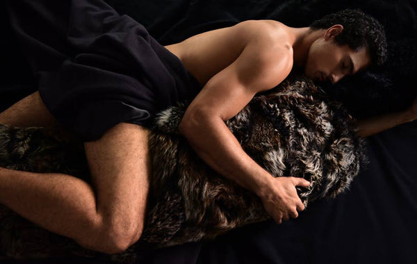 A man in bed with a black bedsheet hugging a huge real truffle brown sheep skin fur body pillow