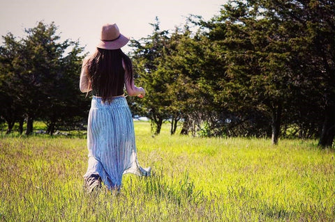 Woman with long brown hair wearing a large felt hat and a long blue skirt, walking through a green field with conifers in the background, at Topsy Farms