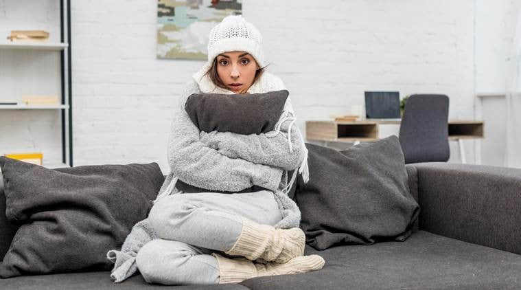 A woman sat curled up on her sofa cuddling a cushion in a hat, scarf, coat, gloves, and wooly socks.