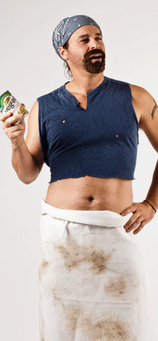 a man with a goatee has a bandana on his head, wears a cropped t-shirt, a dirty apron, and holds a can of vegetables