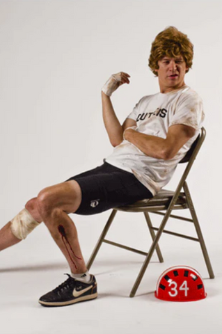 a dirty man with shaggy hair sits on a folding chair and wears a white t-shirt and bike shorts with a bike helmet next to him