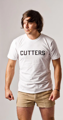 man with shaggy hair wears tan shorts and white t-shirt that says cutters on it