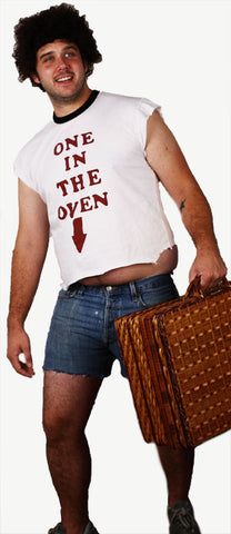 a man wears curly hair, a sleeveless shirt that says one in the oven, short jean shorts, and carries a suitcase