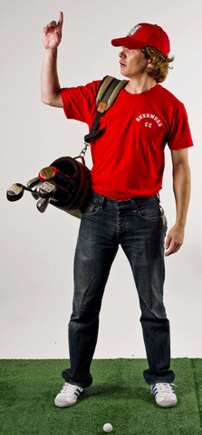 a man points in the air and wears a red cap, red t-shirt, jeans, and carries a golf bag full of clubs