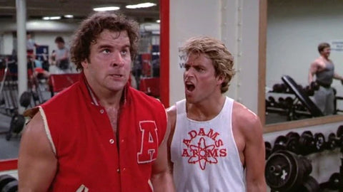 two men are in a weight room, one wears a red letter jacket with the sleeves cut off, one wears a white tank top