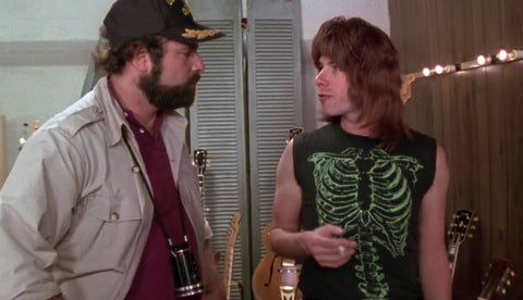 two men stand in a room full of guitars, one wears a t-shirt with a green skeleton on it, and the other a baseball cap, and khaki shirt