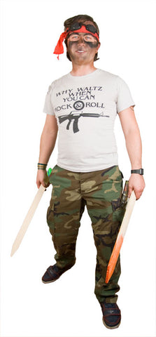 a man with a dirty face wears a red bandana on his head, tan tshirt, camouflage pants, and hold two wooden stakes