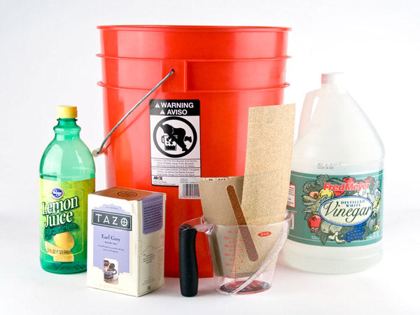 a red plastic bucket, a bottle of lemon juice, a box of earl grey tea, a bottle of vinegar, two samples of sandpaper, a nail file, and a plastic measuring cup