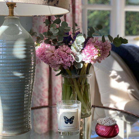 A Collingwood of Somerset Orange Blossom & Fig Candle on a glass table in a country home surrounded by pink hyacinths