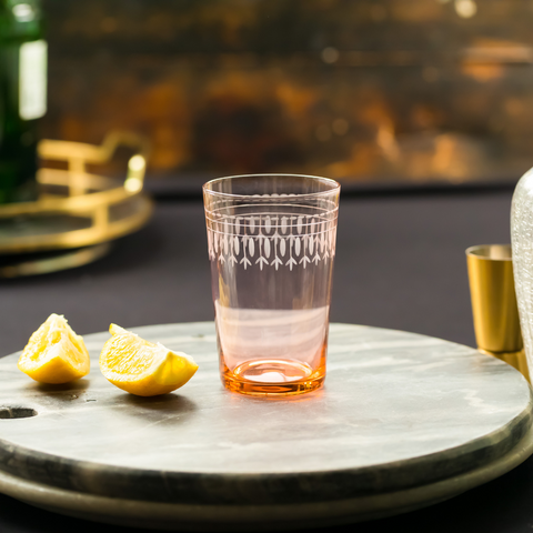 A hand blown and hand engraved rose glass tumbler from The Vintage List on a marble tray with slices of lemon on the side