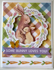 Handmade Easter card with illustrated bunnies hugging inside a floral wreath, set on pastel background