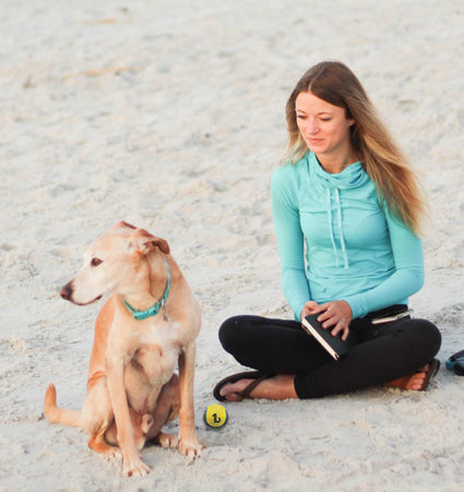 A woman and her dog sitting on a beach with the be one breead strudy tennis ball with them.