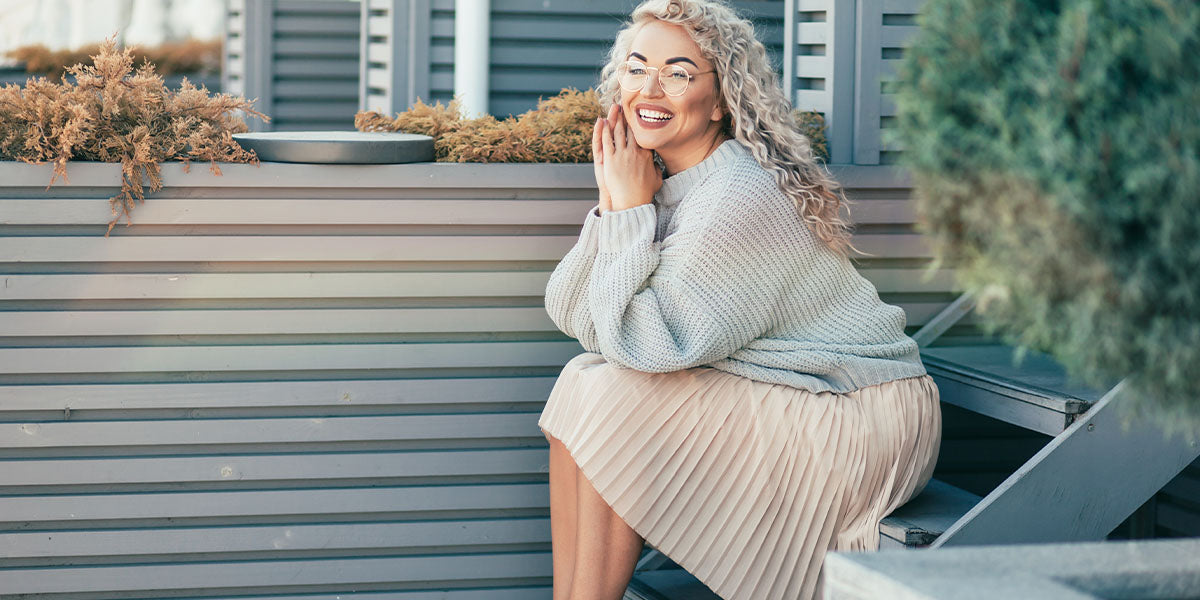 Beautiful plus size model with blond curly hair wearing grey knitted sweater and hipster glasses posing on city street. Fashion everyday outfit for cold season.