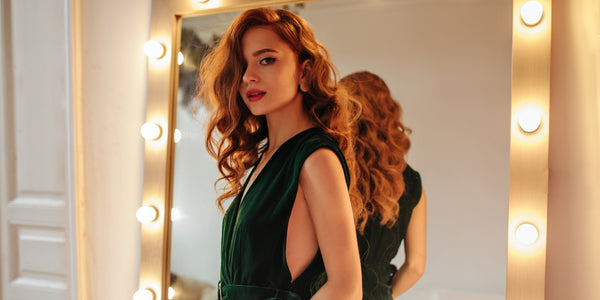 Cheerful woman in dark green outfit poses near mirror. Lovely lady with ginger wavy hair in stylish dress looking into camera.