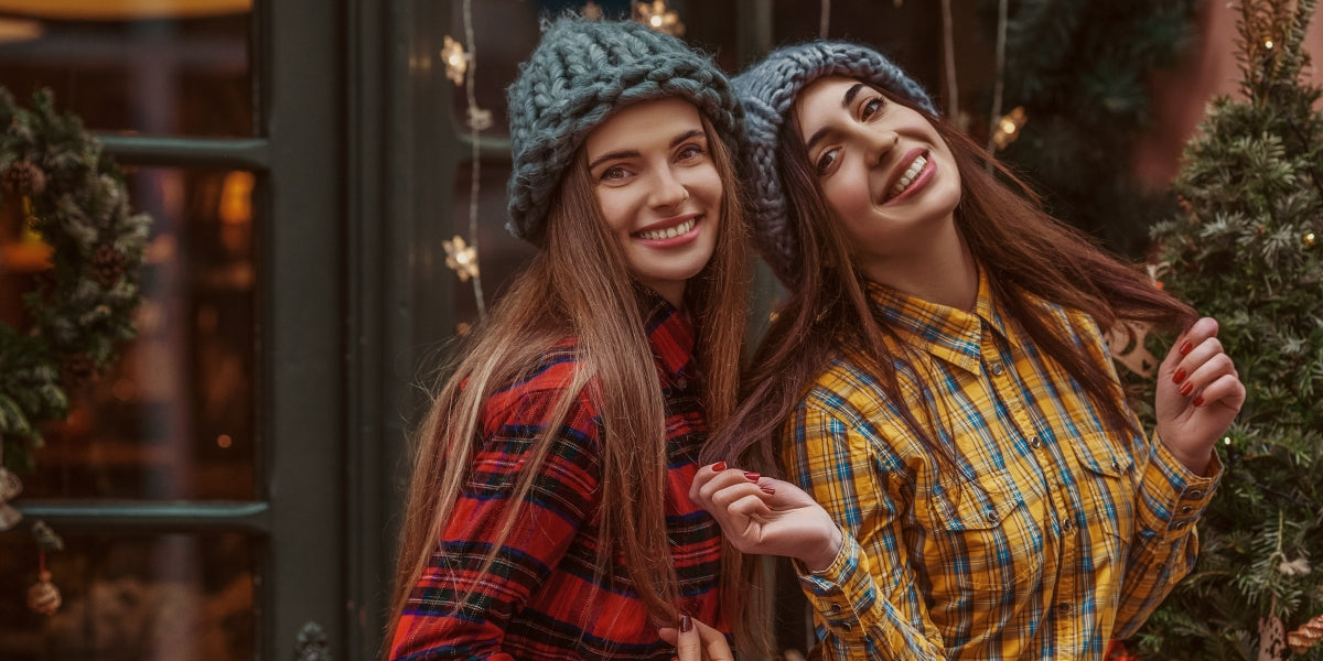 Christmas, New Year, winter holidays conception two happy smiling women, friends, sisters, wearing knitted hat, tartan shirts, posing in street with festive decorations.