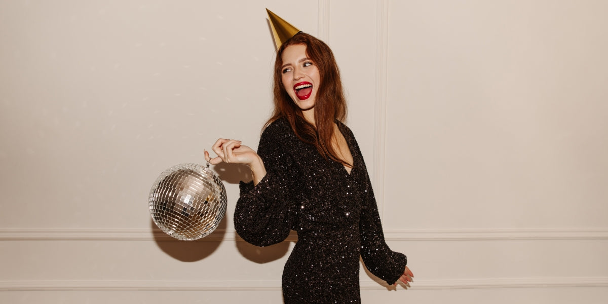 Fashionable young caucasian girl in black dress posing with disco ball looking away on white background. Red-haired lady comes off on holiday alone. Party concepts