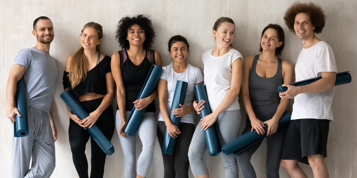Group portrait of happy diverse people with yoga mats in hands, posing for photo at grey wall background, looking at camera, smiling young men and women having fun after training in studio