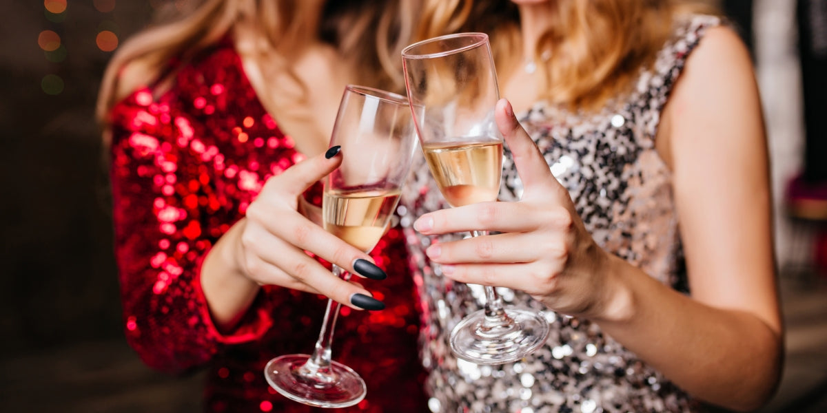 Indoor close-up photo of two beautiful girls wearing New Year's hats celebrating holidays together. Portrait of funny ladies clinking glasses of champagne on Christmas day.