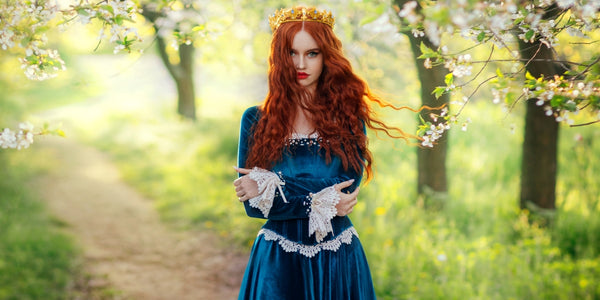 Redhead fantasy woman queen. Blue long velvet dress vintage old style clothing. Red curly hair flying waving in wind. Summer nature green flowering trees garden. Girl Princess beauty face.