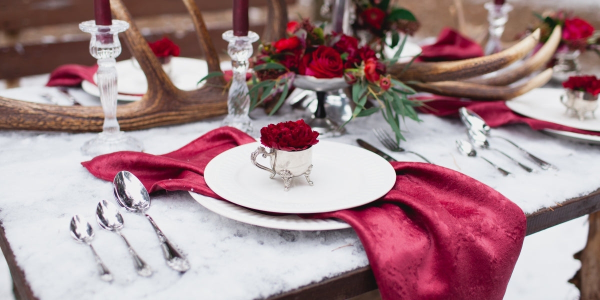 Wedding table setting in masala colors with plates, cutlery, red floral composition, candles, velvet napkins, antlers on the table covered with snow