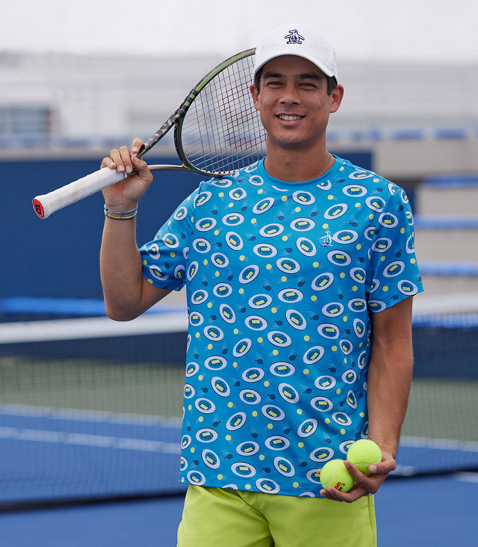 Tennis player wearing an Original Penguin shirt and hat holding a racket and 2 tennis balls