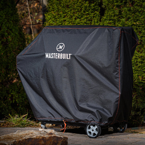 A covered Gravity Series grill sits on an outdoor patio. The cover is black with the Masterbuilt name and logo printed on it in white.
