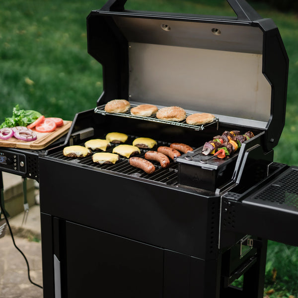 Cheeseburgers and sausages cook on the main cooking grate of an open AutoIgnite grill while hamburger buns toast on the warming rack and kebabs sear on the hopper grill plate. A wooden cutting board holding slices onions, tomatoes and lettuce sits on the left side shelf above the digital control panel. A full drinking glass sits on the right side shelf.