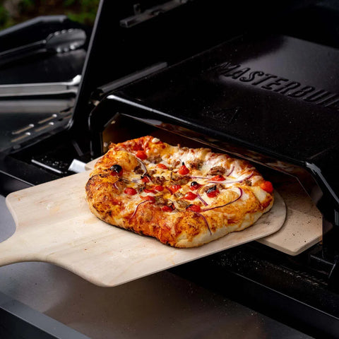 A cooked pizza is pulled from the Masterbuilt Pizza Oven using a wooden peel