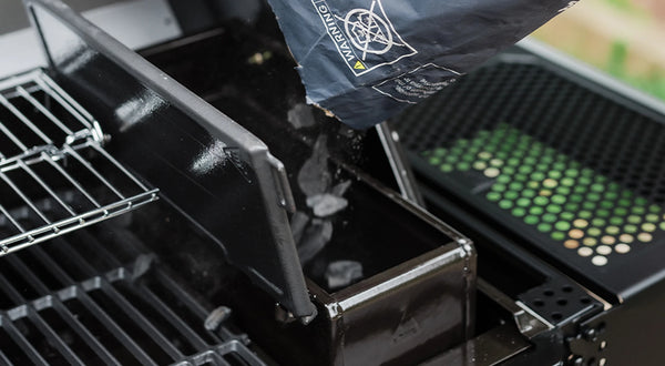 Someone pours lump charcoal from a bag into the hopper of an AutoIgnite grill. Part of the grill grate and warming rack are visible to the left. The right side shelf may be seen to the right of the hopper.