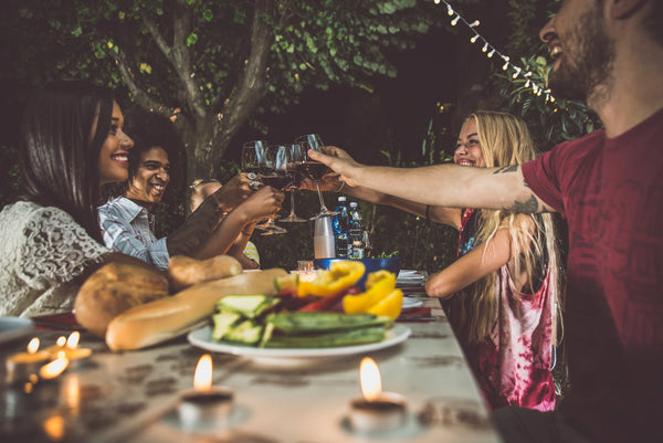 A diverse group of people sit around an outdoor table at night. Candles burn on the table and stings of lights hang from the trees. The table is full of food, and the four friends clink wine glasses in a toast.