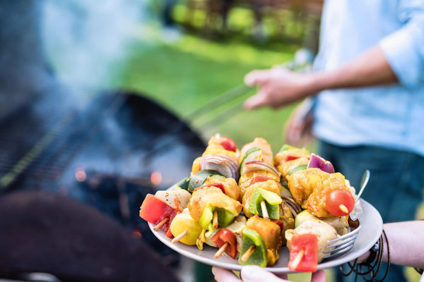 A person holds a plate piled with kebab skewers in both hands. A second person holding long tongs over a lit grill are visible but blurred in the background.