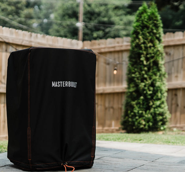 A covered vertical smoker sitting on an open patio. A needle cypress tree and wooden privacy fence are in the background.