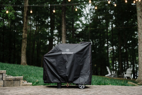 A covered Masterbuilt digital charcoal grill and smoker sits on a paved patio in front of a lawn and trees
