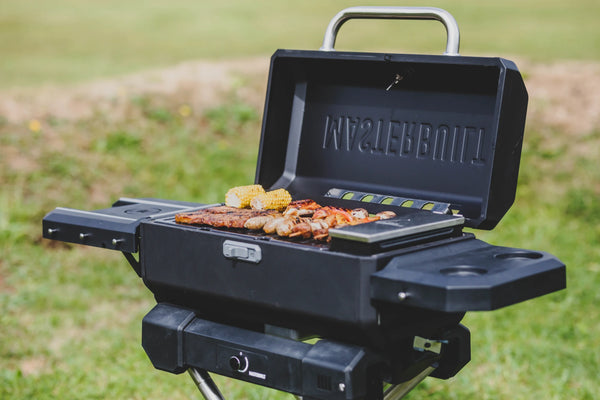 A Masterbuilt portable grill on a QuickConnect cart with 2 side shelves. The side shelf on the right has cupholders. The grill is open to show the charcoal hopper and a variety of food cooking on the grill.