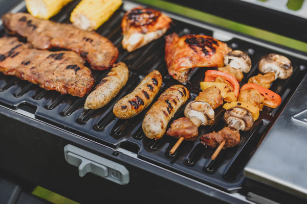 Steaks, sausages, kebab skewers, chicken breasts, and corn on the cob cooking on the grill of a Masterbuilt portable grill. The charcoal mini-hopper is just visible on the right.