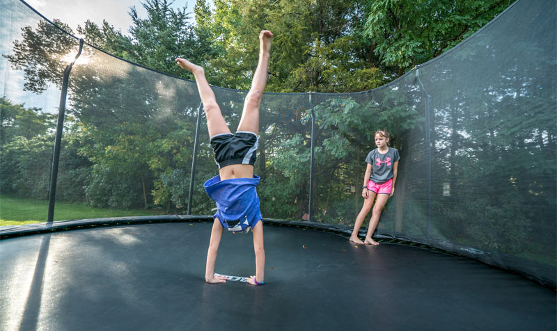 Tyttö seisoo käsillään trampoliinilla ja toinen tyttö nojaa trampoliinin turvaverkkoon