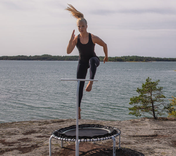 Nainen hyppii fitness trampoliinilla ulkona kalliolla merimaisema taustalla
