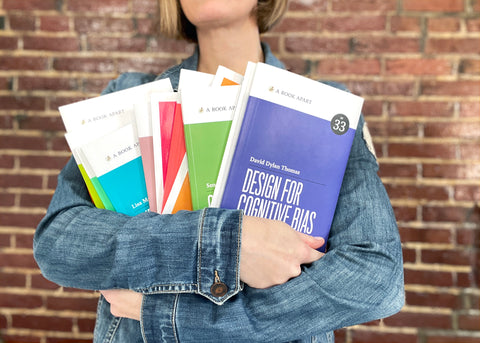 A white woman wearing a jean jacket holds several colorful books in her arms. She stands in front of a brick wall.