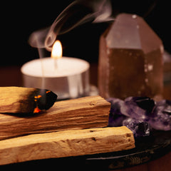 Palo santo burning gently next to a candle and some small crystals, showcasing the smoke and herbs cleansing method for crystals.