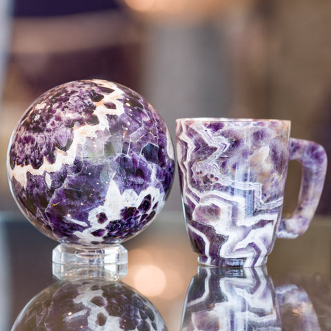 A crystal sphere and teacup both made of chevron amethyst sitting on a glass table