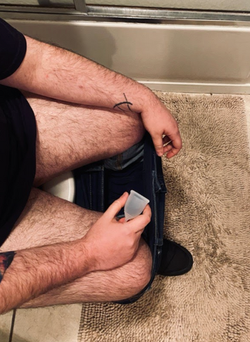 Transman sitting at toilet with pants don holding menstrual cup