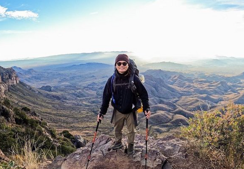 A male The Backpacker employee wearing a backpack and holding trekking poles stands at edge of a cliff with mountain range behind him.