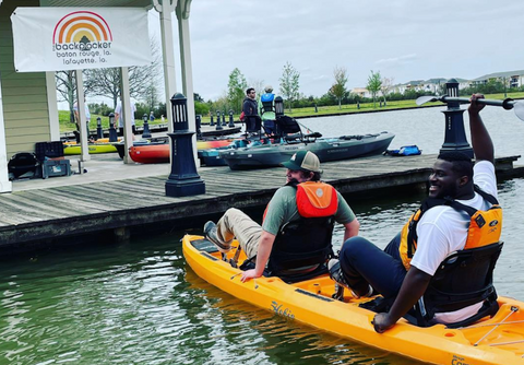 Two male The Backpacker employees sit in a kayak on water near a dock.