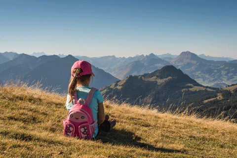 Girl with pink backpack on sitting on the grass looking out over a mountain range