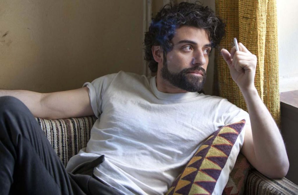 Man with beard and disheveled hair holding a cigarette by the window