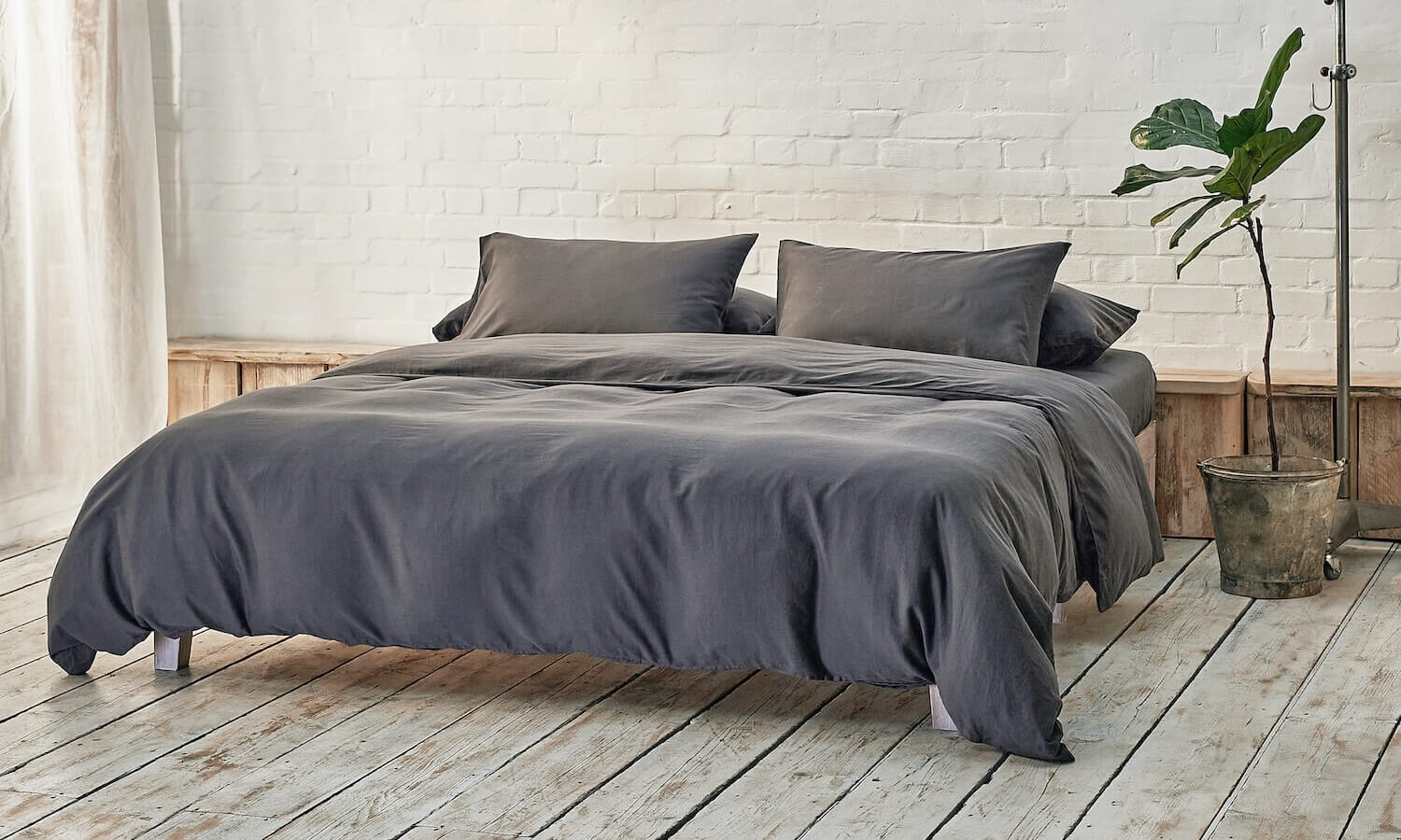 dark grey duvet cover, two pillowcases, and bottom sheet on double bed in apartment with plant and wooden flooring