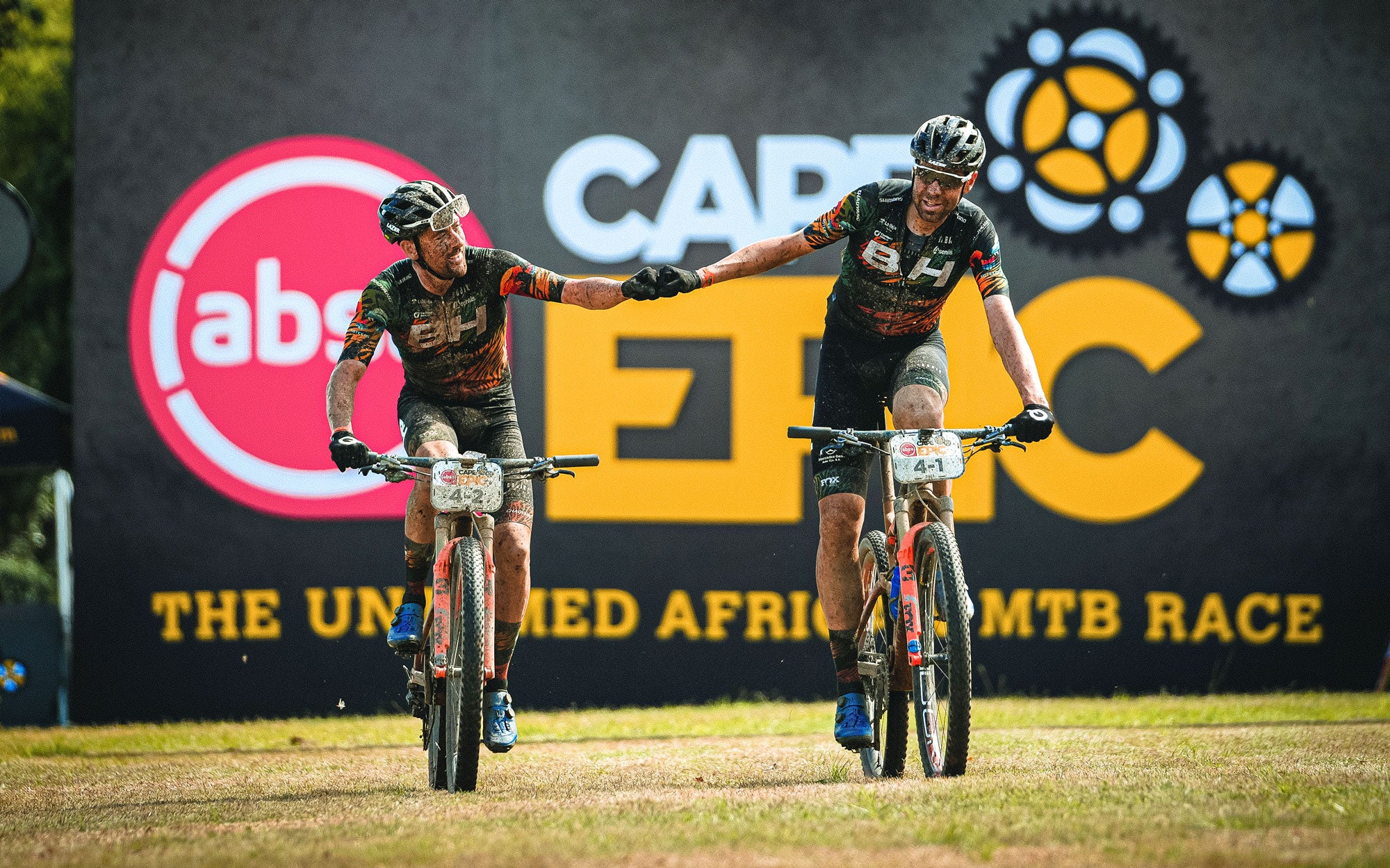 Rodríguez and Valero, from BH Coloma, shake hands as they enter the finish line after finishing a stage of the race. They are covered in mud and their facial expression is a mixture of exhaustion and happiness. 