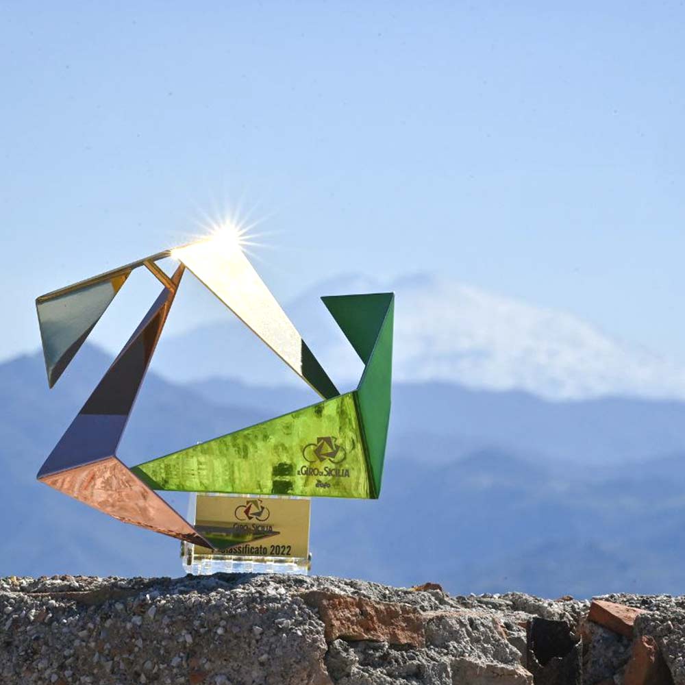 Gedenktrophäe des Giro di Sicilia 2022 auf einem Felsen, hinter dem und durch den hindurch man einen schneebedeckten Berg auf dem Gipfel sehen kann.