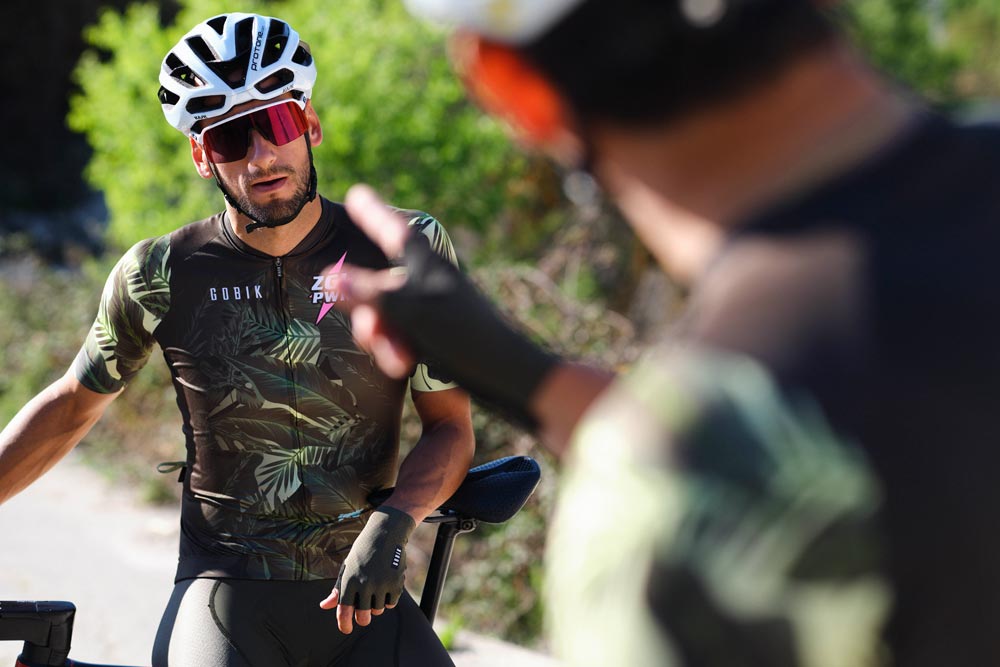 Deux cyclistes discutent en s'appuyant sur leur vélo. De face, le kit de Gobik Custom Services pour l'équipe ZGL Power, avec un design dans des tons verts rappelant des paysages verdoyants, avec le nom et le logo de l'équipe en rose bubblegum.  