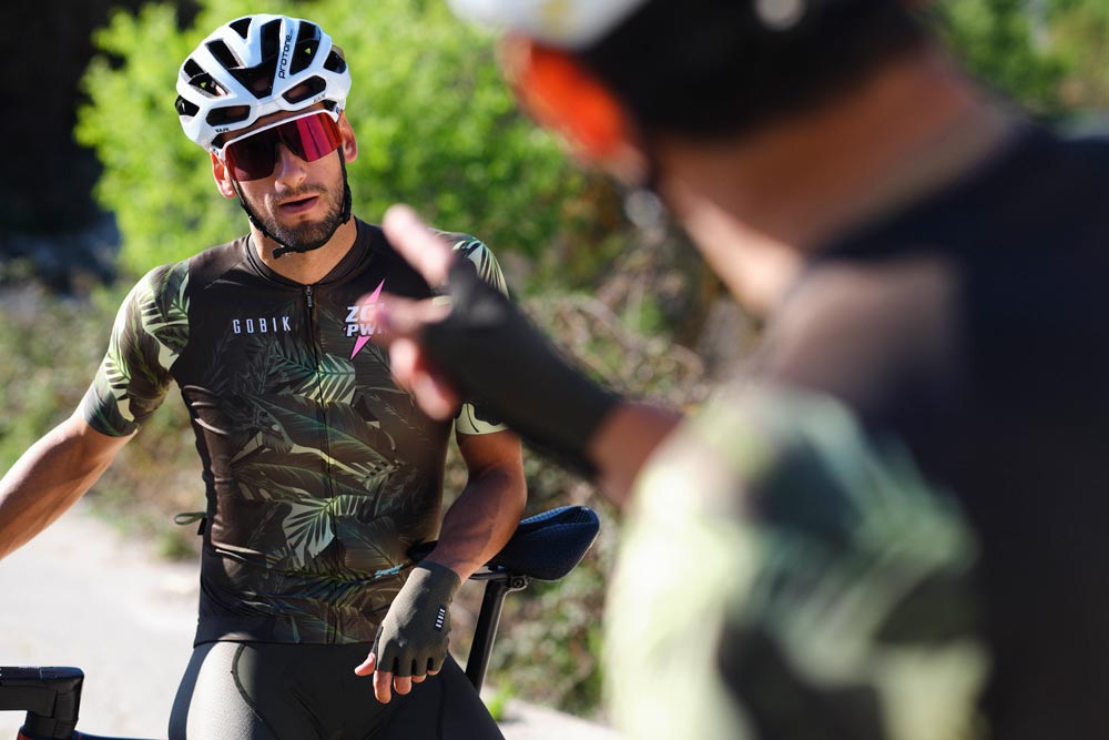 Dos ciclistas teniendo una conversación apoyados en sus bicicletas. De frente, el kit de Gobik Custom Services para el equipo ZGL Power, con un diseño en tonalidades verdes que recuerda a paisajes de gran vegetación, con nombre del equipo y logo en rosa chicle.  